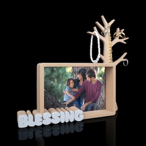 以下が含まれることがあります： ベース部分に白いブロック体で「BLESSING」という文字が刻まれた明るい色の木製フォトフレーム。右側には木のような形をしたジュエリースタンドがあり、白いビーズのネックレスとゴールドの指輪が掛けられています。
