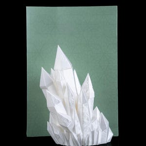 Może przedstawiać: Biała formacja kryształów origami z ostrymi, kątowymi fasetami. Papierowa rzeźba umieszczona jest na zielonym tle z geometrycznym wzorem, tworzącym uderzający kontrast. Dzieło prezentowane jest na odblaskowej czarnej powierzchni.