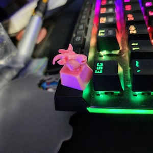 May include: A pink 3D printed keycap with a goat design, sitting on a black keyboard with a green light strip. The keyboard has the word "ESC" on one of the keys.