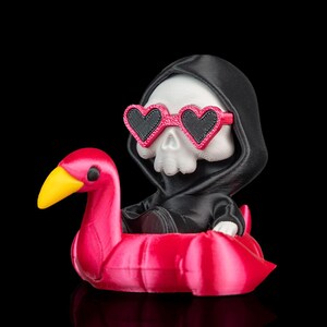 May include: A 3D printed figurine of a white skull wearing a black hooded robe and pink heart-shaped sunglasses. The skull is sitting inside a bright pink inflatable flamingo pool float with a yellow beak and black eyes, set against a black background.