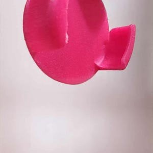 May include: A bright pink, abstract-shaped object with a rounded main body and a rectangular extension. The object appears to be made of a solid material, possibly plastic or resin, and is set against a plain white background.