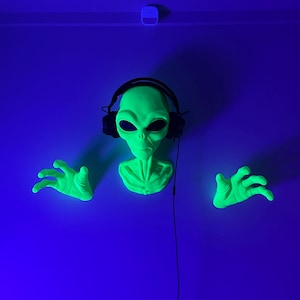 May include: A green alien head with black headphones, two green hands, and a black cord hanging down against a blue background.