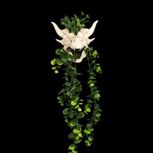 Puede incluir: Una maceta blanca con forma de calavera con cuernos y follaje verde. La maceta tiene una superficie detallada y texturizada y est&aacute; llena de varias plantas verdes, incluyendo eucalipto colgante. El fondo es negro s&oacute;lido.