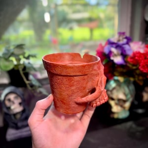 May include: A terracotta-colored skull-shaped planter. The planter has a textured surface and a handle formed from the skull's jaw. The interior is hollow, suitable for small plants or decorative items. The planter is held in a hand.