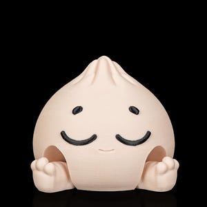 Puede incluir: Una figura redondeada de color beige claro con forma de dumpling al vapor y punta superior. Presenta un rostro sencillo impreso con dos ojos negros curvados y una boca pequeña y sutil. La base tiene dos pequeñas protuberancias redondeadas.