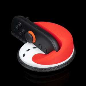 May include: A black and red gaming controller rests on a white and red base. The controller has black buttons and an orange accent. The base has a black, textured border and a curved red design.