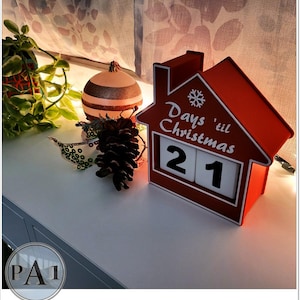 May include: A red house-shaped Christmas countdown calendar displaying "Days 'til Christmas" and the number "21". The calendar sits on a white surface, with a decorative bauble and pine cone in the background.