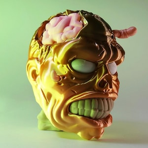 May include: A grotesque, golden-yellow zombie head with an exposed pink brain and a worm protruding from the top. The zombie has green teeth, white eyes, and a menacing expression. The head is likely a Halloween decoration or prop.