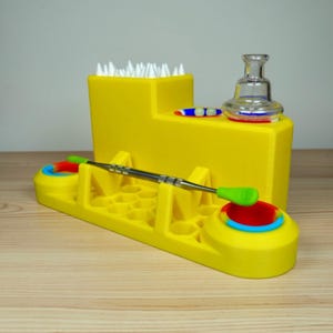 May include: A yellow multi-functional organizer with a variety of compartments and features. It includes a clear glass piece, a metal tool with green tips, and a section filled with white items. The organizer has red, blue, and green accents.