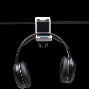 Puede incluir: Auriculares negros sobre la oreja colgando de un soporte blanco y negro. El soporte está montado en una barra negra. Los auriculares tienen detalles plateados sobre un fondo negro.