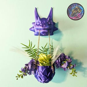 May include: Purple dragon head planter with a hanging pot filled with purple and yellow flowers. The dragon head is 3D printed and has a textured surface. The pot is also 3D printed and has a textured surface.