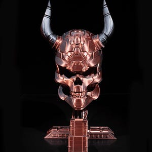 Può includere: Una scultura a forma di teschio color rame con corna argentate, montata su una base abbinata. Il teschio presenta un design meccanico dettagliato, con denti e lineamenti del viso visibili. Le corna si incurvano verso l'alto, aggiungendo all'aspetto imponente della scultura.