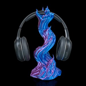 Puede incluir: Unos auriculares negros descansan sobre un soporte para auriculares arremolinado. El soporte es una mezcla de tonos azules y morados, con un diseño en forma de llama. El fondo es negro, resaltando el soporte y los auriculares.