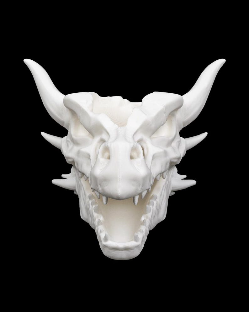 Puede incluir: Escultura de cr&aacute;neo de drag&oacute;n blanco con cuernos, dientes afilados y boca abierta. La escultura tiene una superficie texturizada y detallada, sobre un fondo negro. Es un art&iacute;culo decorativo de tem&aacute;tica fant&aacute;stica.