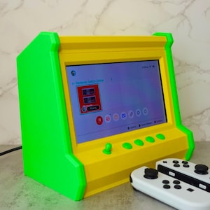 May include: A green and yellow arcade-style console with a screen displaying the Nintendo Switch home screen. Two white Nintendo Switch Joy-Con controllers are on the table next to the console.