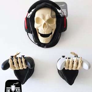 Può includere: Un supporto per cuffie a forma di teschio con cappuccio nero e cuffie rosse e nere. Sotto, due supporti per controller, uno nero e uno bianco, ciascuno con un design a mano scheletrica. Il logo "IK 3D" è in basso.