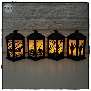 Puede incluir: Cuatro linternas negras de Halloween con interiores naranjas iluminados. Cada linterna presenta un diseño espeluznante diferente: murciélagos, calabazas, una escena de cementerio y manos de zombis. Las linternas son rectangulares con una parte superior decorativa.