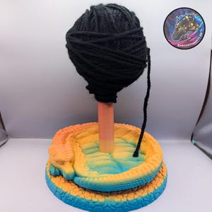 Peut inclure: Un support à fil avec une pelote de laine noire sur un poteau pêche, posé sur une base colorée en forme de serpent. La base passe du bleu au jaune, avec une tête de serpent détaillée. Un logo avec le texte "Kekreationsart 3D" est visible.