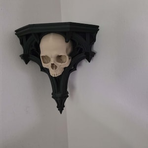 May include: A black wall-mounted corner shelf featuring an intricate, pointed architectural frame. Centered on the front of the shelf is a realistic, off-white human skull sculpture integrated into the design, providing a decorative focal point.