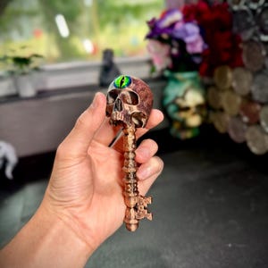 Può includere: Una chiave color bronzo con una testa a forma di teschio e un design ornato dettagliato. Il teschio ha un occhio verde e oro. La chiave è tenuta in una mano, con uno sfondo sfocato con fiori e una finestra.