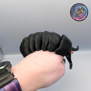 May include: A black, segmented, 3D-printed articulated creature, possibly a centipede or similar insect, held in a hand. The creature has a textured surface and is posed with its legs extended. A circular sticker with a stylized horse head logo is in the background.