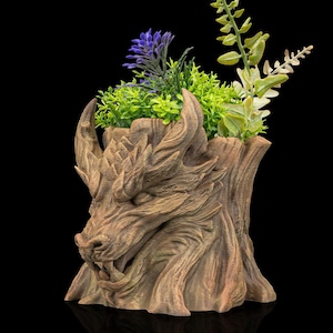 Pode incluir: Um vaso marrom em forma de dragão com um design detalhado e texturizado. O vaso é preenchido com uma variedade de plantas artificiais, incluindo folhagens verdes e roxas. O vaso é feito de um material semelhante à madeira e fica em um fundo preto.