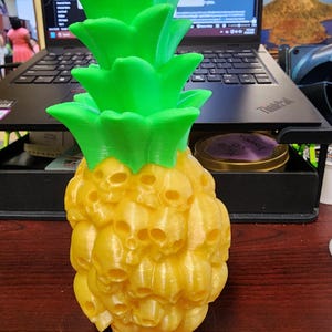 May include: A 3D-printed pineapple sculpture with a unique design. The pineapple body is yellow and features a pattern of skulls, while the top is bright green. This decorative item is a creative blend of tropical fruit and gothic imagery.