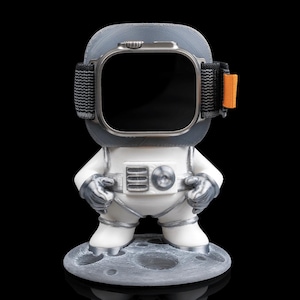 Könnte beinhalten: Eine weiße Astronautenfigur mit einer silbernen Uhr auf dem Kopf, die auf einem grauen mondförmigen Sockel steht.
