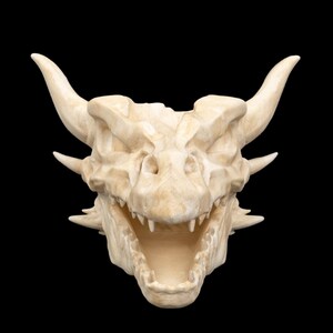 Puede incluir: Un cr&aacute;neo de drag&oacute;n blanquecino con dientes afilados y cuernos prominentes, sobre un fondo negro. La estructura &oacute;sea detallada y la boca abierta sugieren un tema de fantas&iacute;a. Art&iacute;culo decorativo.