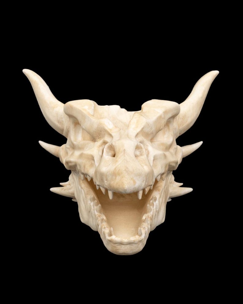 Puede incluir: Un cr&aacute;neo de drag&oacute;n blanquecino con dientes afilados y cuernos prominentes, sobre un fondo negro. La estructura &oacute;sea detallada y la boca abierta sugieren un tema de fantas&iacute;a. Art&iacute;culo decorativo.