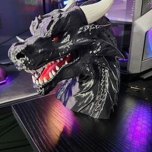 Puede incluir: Una escultura de cabeza de dragón negra con cuernos blancos, ojos rojos y una boca abierta detallada que revela dientes blancos y un interior rojo. La escultura está sobre una superficie negra.