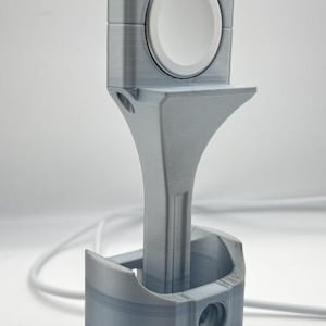 May include: A gray 3D-printed Apple Watch charging stand. The stand is designed to resemble an engine piston and connecting rod. The watch sits in a cradle at the top, with the charging cable extending from the base. The stand is approximately 5 inches tall.