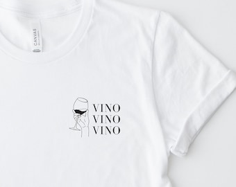 May include: A white short-sleeve t-shirt featuring a black line art graphic on the left chest. The graphic depicts a hand holding a wine glass with the text VINO VINO VINO stacked vertically to the right of the illustration.