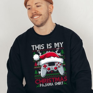 Op de afbeelding: Zwart sweatshirt met een feestelijk ontwerp met een kerstmuts over een gamecontroller. De tekst "THIS IS MY CHRISTMAS PAJAMA SHIRT" staat in rood en groen.