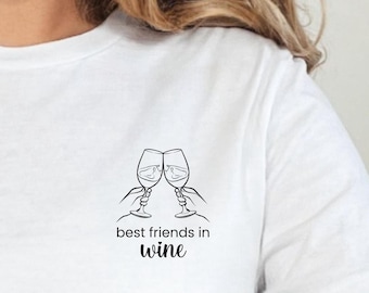 May include: A white short-sleeved t-shirt featuring a black line art graphic on the chest. The design depicts two hands holding wine glasses in a toast, with the text "best friends in wine" printed in a mix of sans-serif and script fonts.