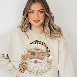 Può includere: Una felpa girocollo color sabbia con una stampa di Babbo Natale che indossa occhiali e un cappello con stampa leopardata. Babbo Natale fa l'occhiolino, ha una barba bianca testurizzata e tiene in mano un oggetto a forma di cuore con glitter.