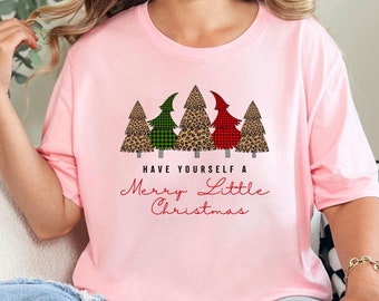 Könnte beinhalten: Ein hellrosa T-Shirt mit kurzen Ärmeln und einer Grafik von fünf stilisierten Weihnachtsbäumen mit Leopardenmuster sowie grünem und rotem Schottenkaro. Unter den Bäumen steht der Text: HAVE YOURSELF A Merry Little Christmas.