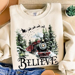 May include: Cream-coloured sweatshirt with a Christmas train graphic. The design includes a red engine, snowy trees, and Santa's sleigh. The word "BELIEVE" is printed in a decorative font. A festive holiday-themed garment.