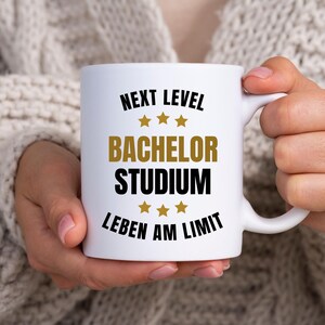 Puede incluir: Una taza de cerámica blanca con texto en negro y dorado que dice NEXT LEVEL, BACHELOR STUDIUM y LEBEN AM LIMIT, decorada con seis estrellas doradas dispuestas en dos filas de tres sobre y debajo del texto central, con un asa lateral.