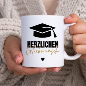 Peut inclure: Tasse en céramique blanche avec un dessin de toque noire et l'inscription allemande "HERZLICHEN Glückwunsch" en noir et or. Un petit cœur noir est en dessous. La tasse est tenue par une personne.