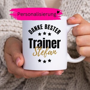 Puede incluir: Una taza de cerámica blanca con texto negro que dice "DANKE BESTER" sobre la palabra "Trainer" y el nombre "Stefan" en letra dorada, con tres estrellas negras arriba y abajo del texto y una banda rosa con la etiqueta "Personalisierung".