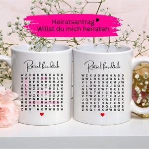 May include: Two white ceramic mugs featuring a word search puzzle with the German text "Rätsel für dich" at the top. The left mug highlights the words "DU MICH HEIRATEN" in pink, while both mugs feature a small red heart icon at the bottom.