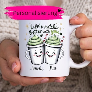 Könnte beinhalten: Ein weißer Keramikbecher mit einer Grafik von zwei lächelnden Tassen, die mit grünem Matcha-Schaum bedeckt sind. Der Text lautet Life's matcha Better with you, darunter stehen die Namen Amelie und Mia. Das Design enthält goldene Sterne und rot gestreifte Strohhalme.