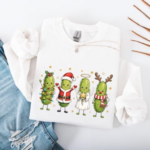 Könnte beinhalten: Ein weißes Sweatshirt mit Rundhalsausschnitt und einem Aufdruck von vier grünen Gewürzgurken in Weihnachtskostümen. Eine Gurke ist mit Lichterketten umwickelt, eine trägt ein Weihnachtsmannkostüm, eine ist als Engel mit Flügeln dargestellt und eine hat ein Rentiergeweih.