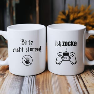 Könnte beinhalten: Zwei weiße Keramikbecher. Auf einem Becher steht "Bitte nicht stören!" mit einem Handsymbol. Der andere Becher hat den Text "Ich zocke" mit einer Spielcontroller-Grafik. Beide Becher haben einen weißen Griff.
