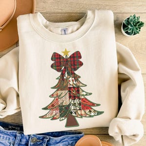 Könnte beinhalten: Cremefarbenes Sweatshirt mit einem Weihnachtsbaum-Design. Der Baum besteht aus Patchwork-Mustern in Rot, Grün und Weiß, gekrönt von einer karierten Schleife und einem goldenen Stern. Ein lässiges, festliches Kleidungsstück.