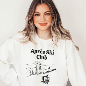 Könnte beinhalten: Ein weißes Sweatshirt mit Rundhalsausschnitt und dem schwarzen Schriftzug "Après Ski Club" über einer schwarzen Strichzeichnung eines Skilifts, Berggipfeln, Tannenbäumen und einer Tasse mit einer Zimtstange und einer Orangenscheibe.