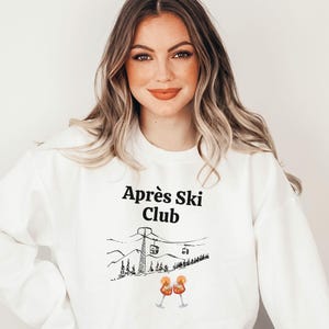 Könnte beinhalten: Weißes Sweatshirt mit dem Text "Après Ski Club" in Schwarz, zusammen mit einer Grafik einer Seilbahn, Bergen und zwei Cocktails. Ein lässiger, bequemer Stil.