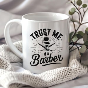 May include: White ceramic mug with the text "TRUST ME I'M A Barber" in black. The design includes a barber chair and crossed scissors. The mug has a curved handle and is set against a soft, textured background.