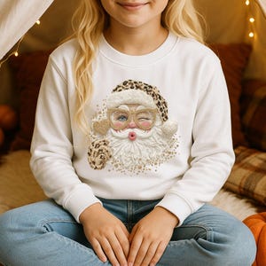 Könnte beinhalten: Ein weißes Langarm-Sweatshirt mit einer Grafik eines zwinkernden Weihnachtsmanns, der eine goldgeränderte Brille und eine Mütze mit Leopardenmuster trägt. Die Grafik zeigt einen strukturierten weißen Bart, ein goldenes Herz und goldene Glitzerakzente.